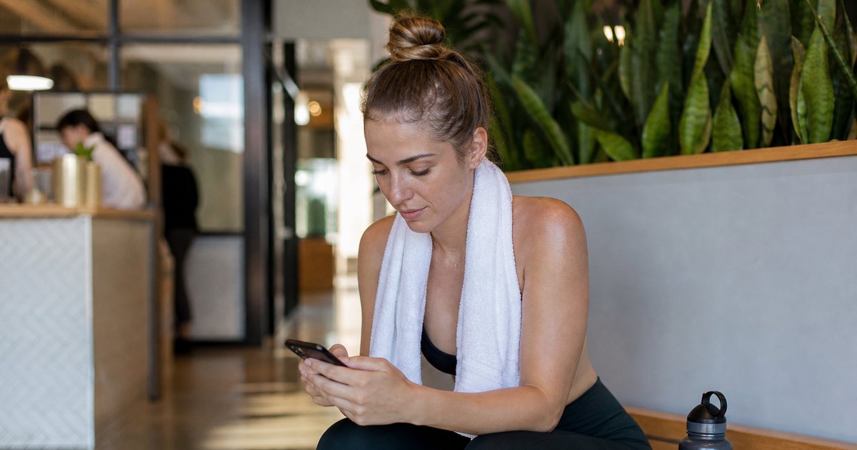 A woman in workout clothes checking her phone while sitting on a bench in a studio lobby after class, towel around her neck, water bottle beside her, warm afternoon light, phone screen not visible