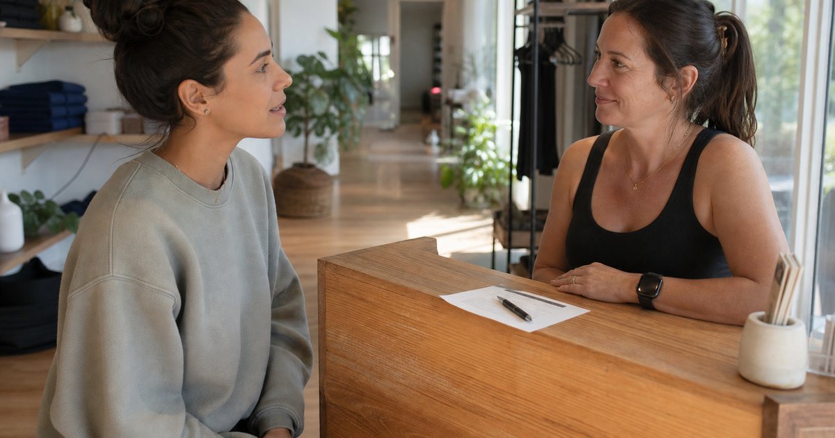 A studio owner and a member sitting at a small wooden front desk in a bright yoga studio, calm conversation, a paper receipt and a pen on the counter, morning light through large windows